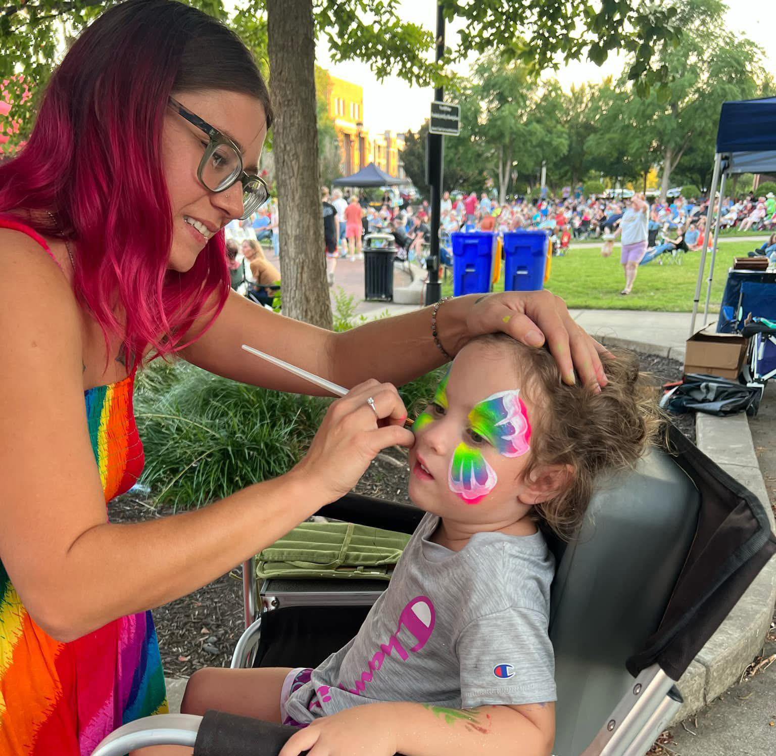Sadie painting a colorful butterfly on a child's face at an outdoor festival